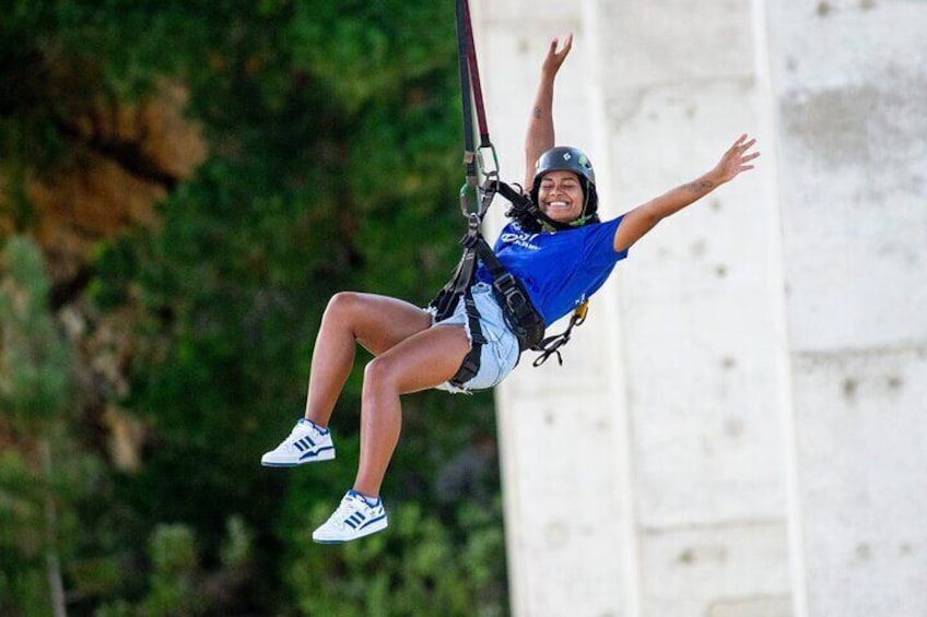 Une personne pratiquant le saut à l’élastique, les bras tendus, portant un casque et un harnais, sur fond de verdure et d’une structure blanche. 5