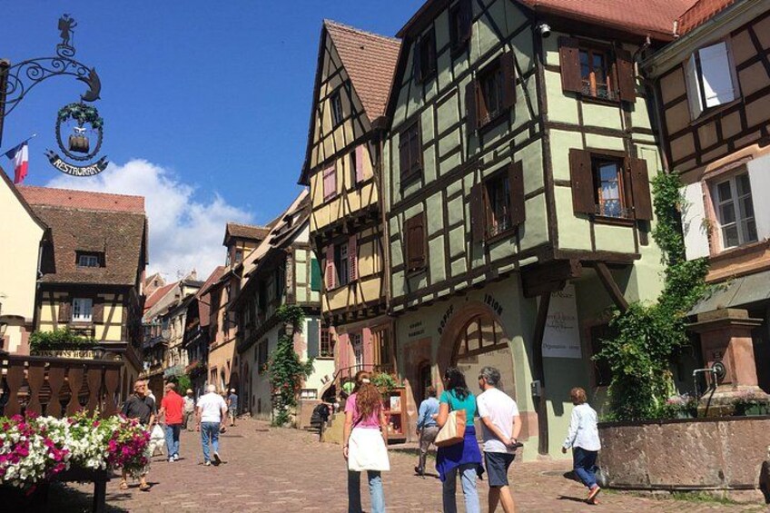 A cobblestone street lined with traditional half-timbered buildings, a sign with a coat of arms, and pedestrians walking. 2
