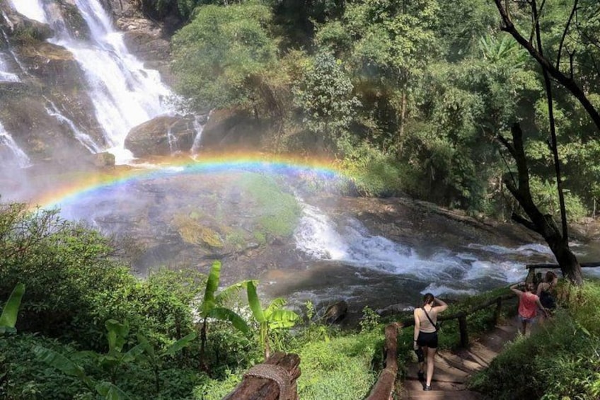 A waterfall with a rainbow, surrounded by lush greenery and a wooden pathway. 1