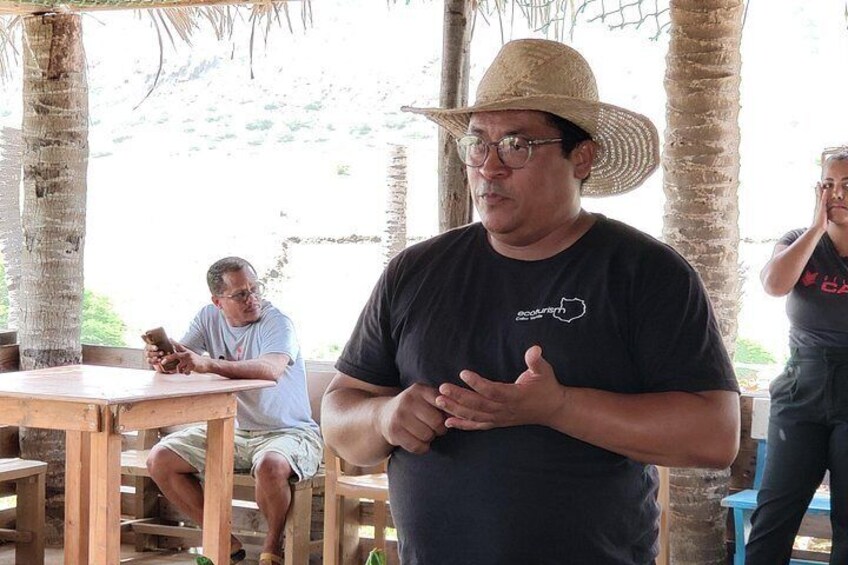 A man in a straw hat and black t-shirt standing in an outdoor setting with palm trees and wooden tables. 3