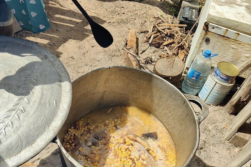 A large metal pot with corn kernels and liquid, a spoon, and a water bottle in a sandy outdoor setting. 1
