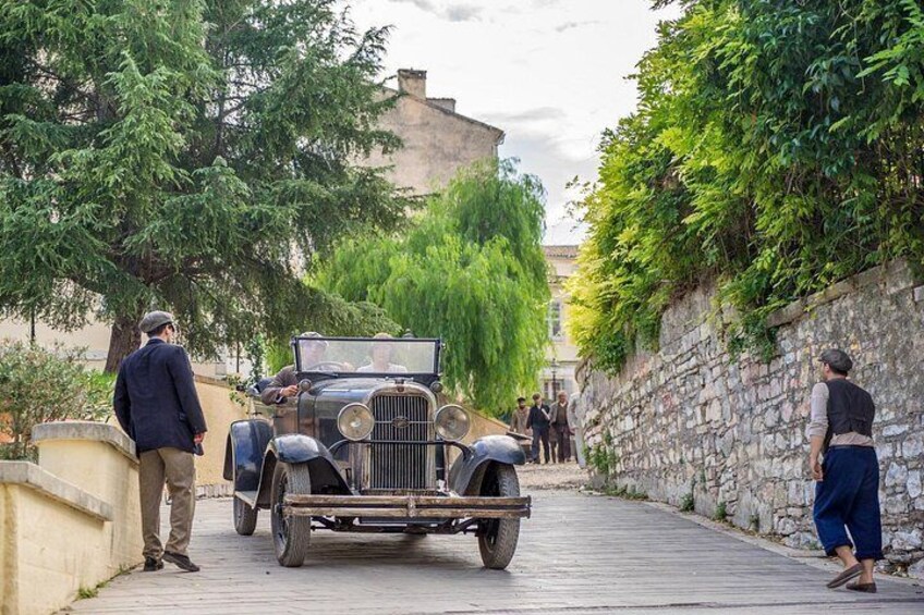 A vintage car parked on a cobblestone street, surrounded by greenery and stone walls. 1