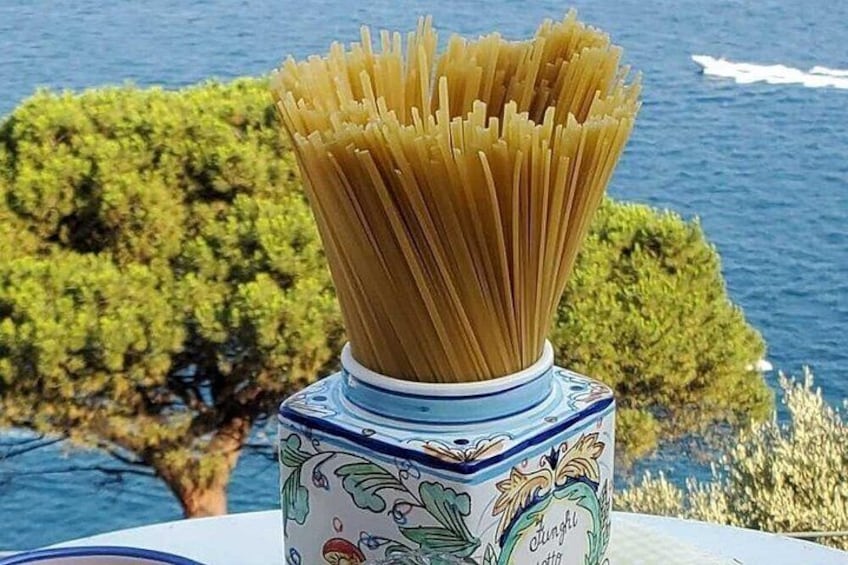 Een bak ongekookte spaghetti met een decoratief deksel, geplaatst op een tafel met uitzicht op de zee en bomen in de achtergrond. 4