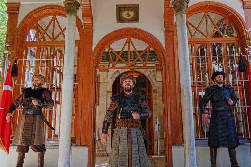 Tres figuras históricas vestidas con trajes de época frente a un edificio con ventanales arqueados y columnas. 2