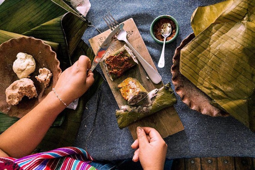 Una persona desenvolviendo comida envuelta en hoja de plátano, junto a un tazón de salsa y un plato de comida sobre una mesa. 1