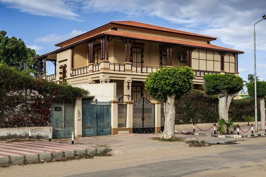 A large, two-story building with a red roof and beige walls, surrounded by a gated area and trimmed trees. 2