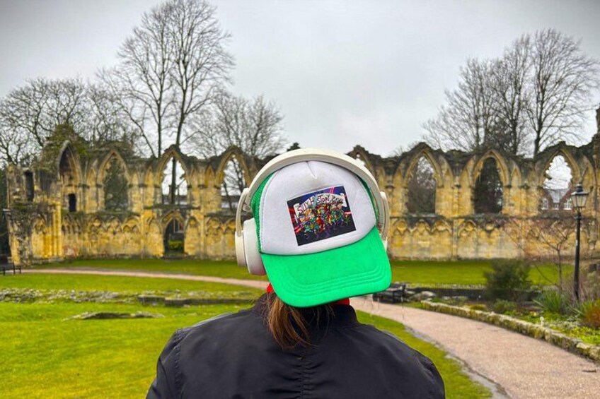 A person wearing a green and white cap and headphones, standing in front of a historic stone building with arches. 5