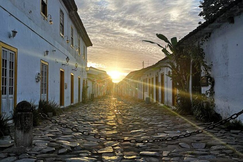 A cobblestone street lined with white buildings, a sunset in the background, and a chain-link fence on the right. 2