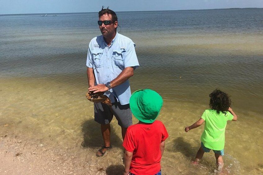 Ein Mann hält einen Krabben auf einem Strand, während zwei Kinder danebenstehen. 3