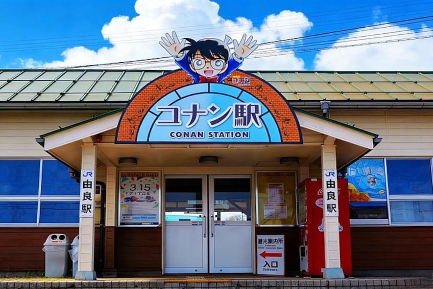 Un edificio con un letrero que dice 'CONAN STATION' y un personaje de caricatura. Hay dos basureros y una puerta con un letrero que indica a la derecha. 1
