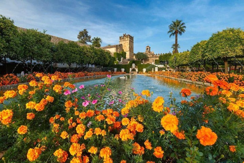 Un jardín con una fuente rodeada de flores de naranja, una estructura tipo castillo y palmeras. 3