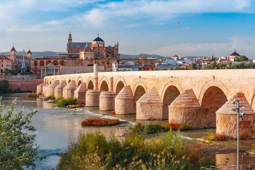 Un puente de piedra con arcos sobre un río, un edificio histórico al fondo y un cielo despejado. 1