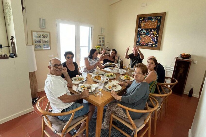 Un groupe de personnes est assis autour d’une table à manger en bois, dans une pièce dont le sol est recouvert de carreaux, qui possède une fenêtre et un tableau accroché au mur. 1