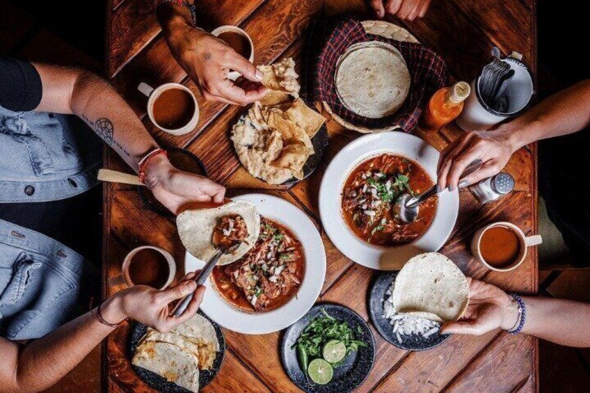 Un grupo de personas cenando alrededor de una mesa de madera con varios platos, incluyendo tazones de sopa y fuentes de comida, además de vasos con bebidas. 1