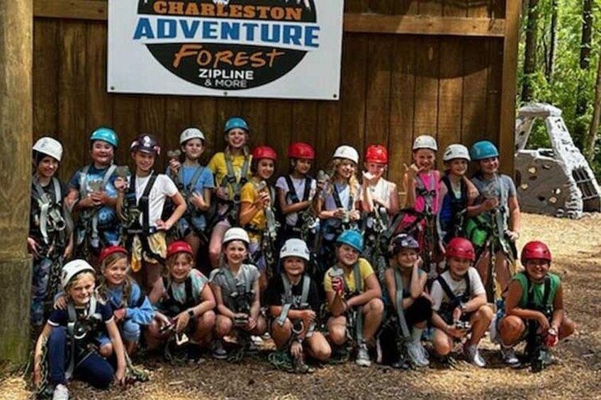 A group of people in helmets and harnesses in front of a sign that reads 'Charleston Adventure Forest Zipline & More'. 2