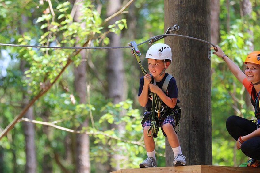 A person in a helmet and harness on a zip line, another person assisting, and a forested area. 1