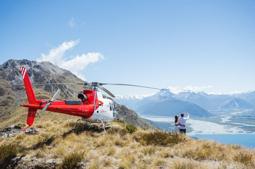 A red and white helicopter parked on a grassy area with a mountainous backdrop and a glacier in the distance. 3