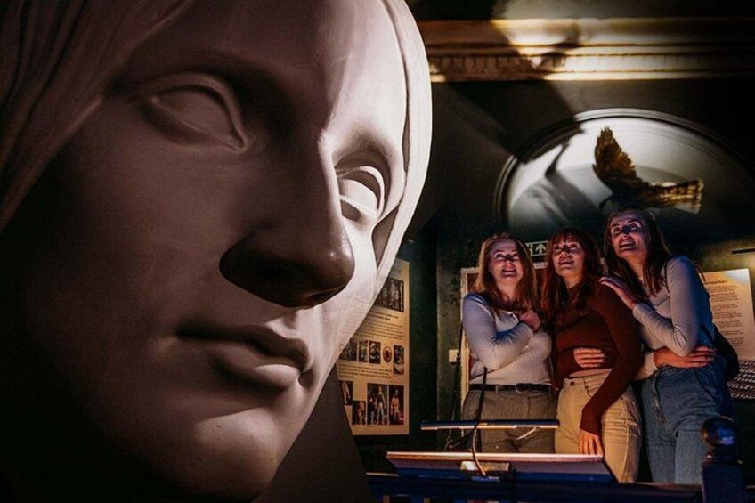 Un busto scolpito, una finestra circolare e tre persone in una stanza con manifesti informativi. 1