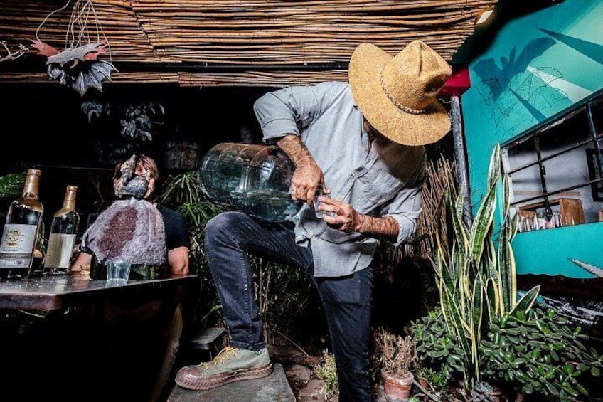 Una persona sirve una copa grande de líquido, posiblemente vino, en un entorno al aire libre con plantas y botellas. 2