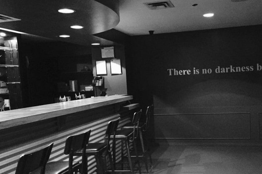 Un bar avec un comptoir et plusieurs chaises. Un mur porte une inscription incomplète qui dit : « Il n’y a pas d  ’obscurité b ». 1