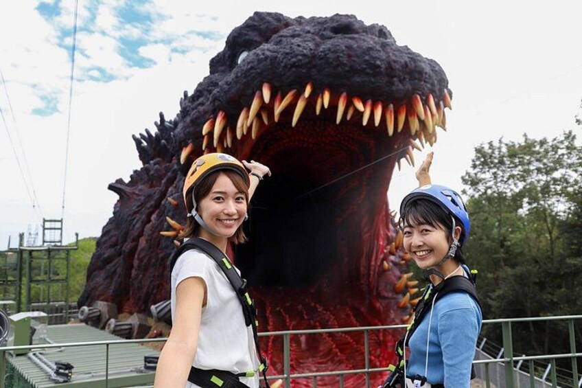 Two people wearing helmets and harnesses in front of a large, menacing creature with sharp teeth. 1