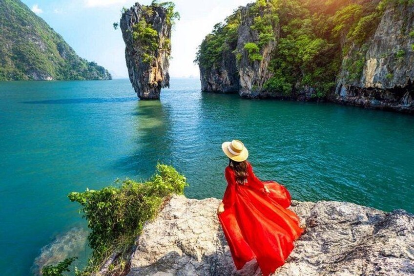 A person in a red dress and straw hat sitting on a rock by a calm sea with a large rock formation in the distance. 1