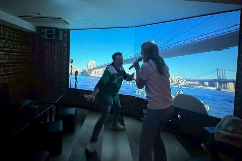 Dos personas cantando karaoke en una habitación con una pantalla grande que muestra un puente. 2