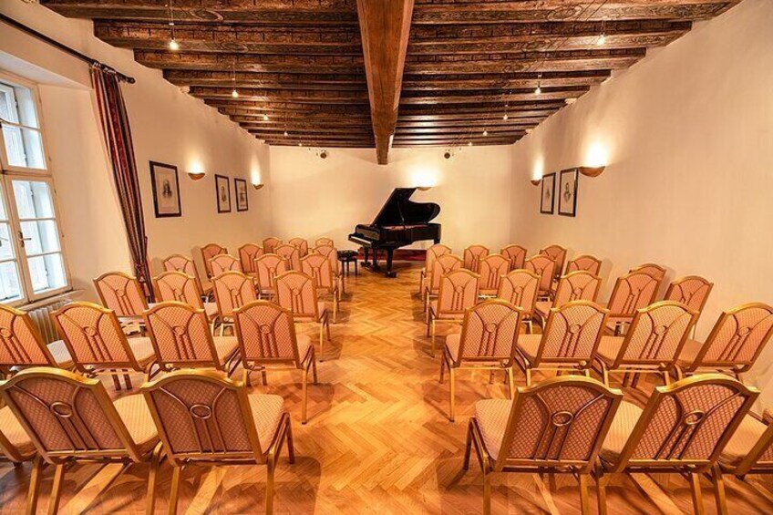 Una habitación con sillas de madera dispuestas en filas, un piano de cola y cuadros enmarcados en las paredes. 3