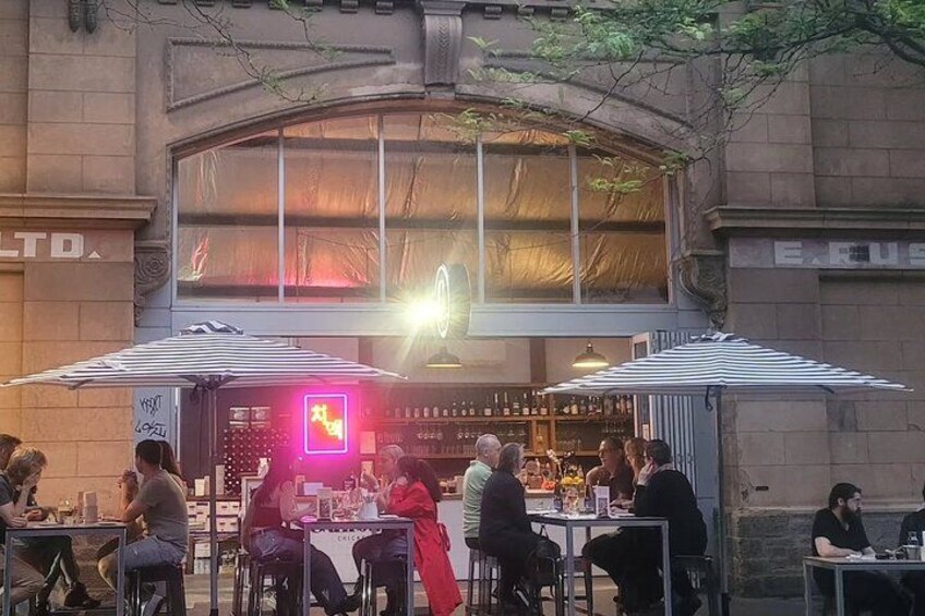 An outdoor dining area with tables and umbrellas, a bar with bottles, and a neon sign. 1