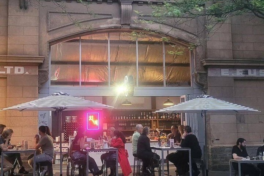 Un espace de restauration en plein air avec des tables et des parasols, un bar avec des bouteilles et un panneau lumineux au néon. 1