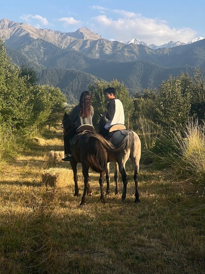 Two people on horseback in a grassy field with mountains in the background. 4