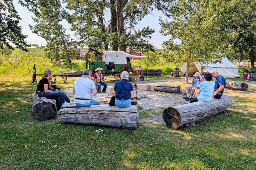 Mensen zitten op boomstammen in een grasrijk gebied, met bomen en een caravan op de achtergrond. 2