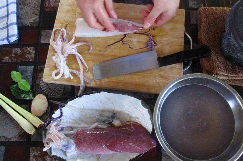 A person cutting meat on a wooden cutting board with a large knife, surrounded by various ingredients and a pot. 2