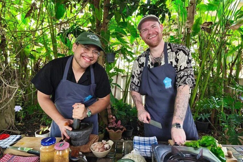 Two people in aprons, one chopping vegetables and the other holding a knife, with a table full of ingredients and a mortar and pestle. 1