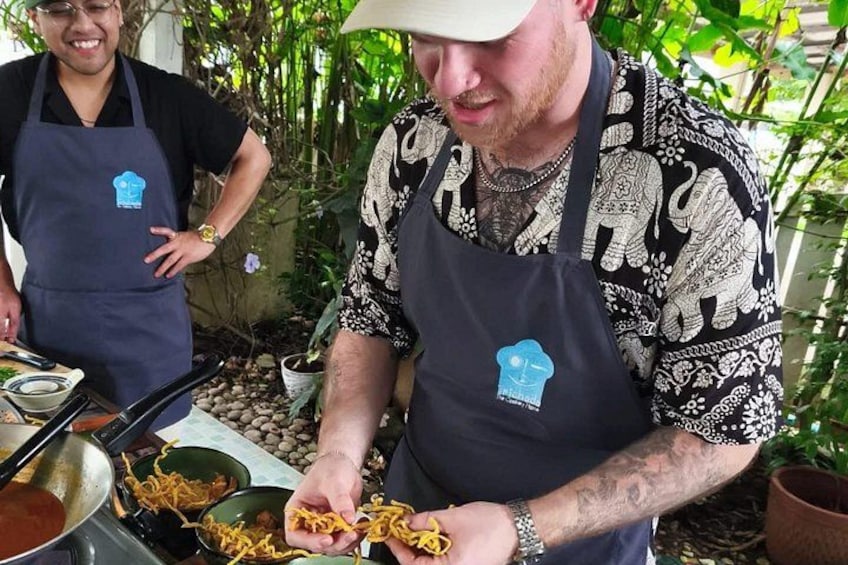 Two people preparing food outdoors, wearing aprons with logos, surrounded by plants and cooking utensils. 5