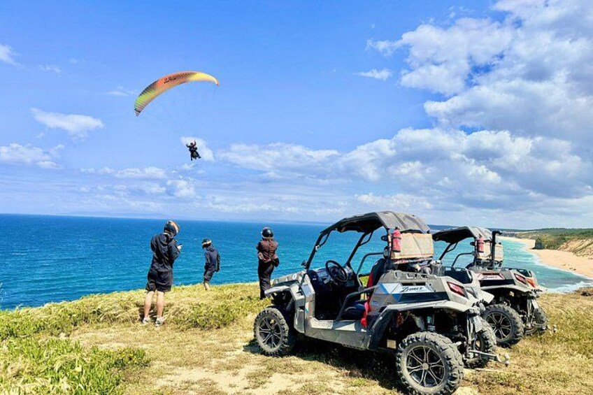 Une plage avec des véhicules tout-terrain, une personne pratiquant le parapente et un ciel bleu dégagé. 1