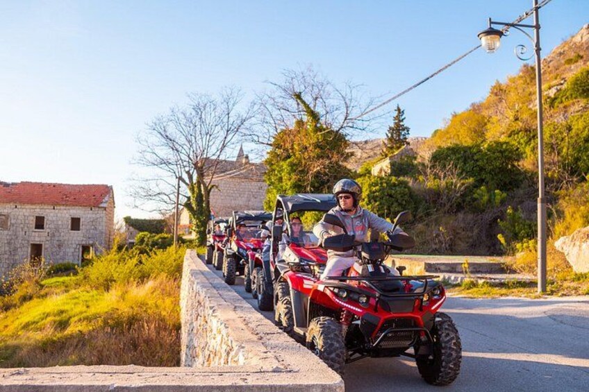 A person riding a red ATV on a paved road with stone buildings and trees in the background. 3