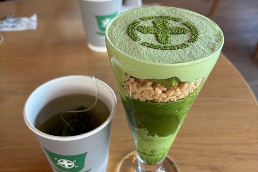 A tall glass with a green beverage, a cup of tea, and a green logo on a table. 5