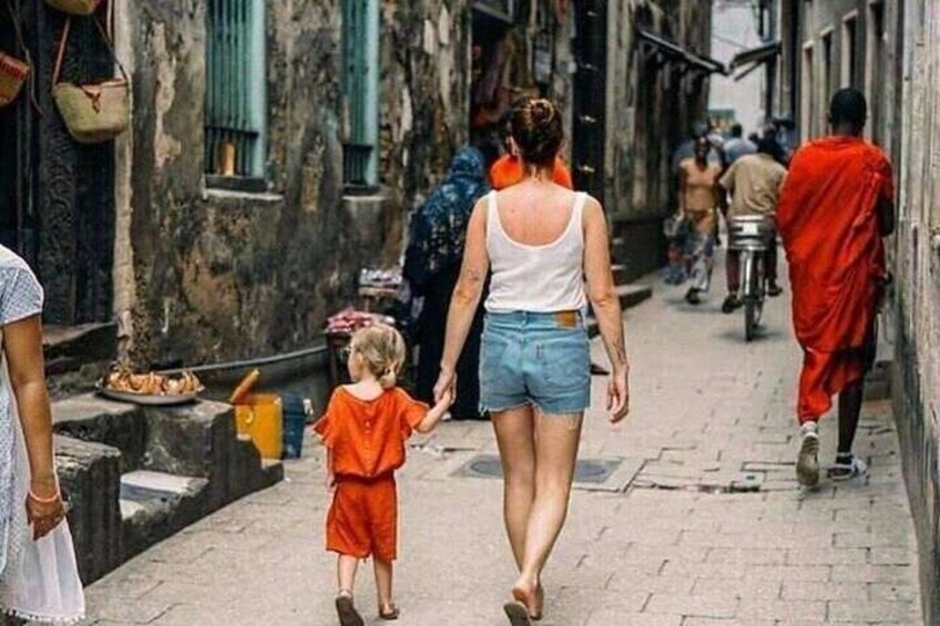 Una stretta via con persone che camminano, un bambino con un completo arancione e una donna con una maglietta bianca e pantaloncini di jeans. 3