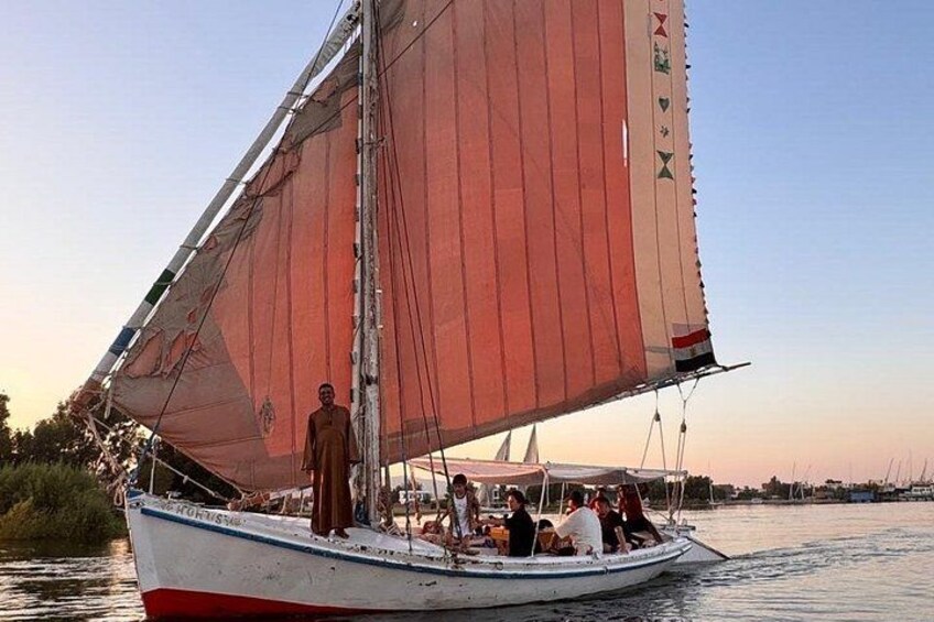 A sailboat with a red sail on calm water, a person standing at the stern, and a few passengers. 5