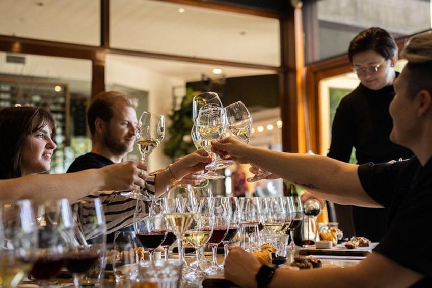 A group of people toasting with wine glasses at a table with various bottles and glasses. 4