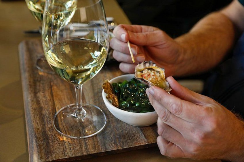 A person holding a piece of food with a toothpick, a glass of white wine, and a bowl of greens on a wooden table. 2