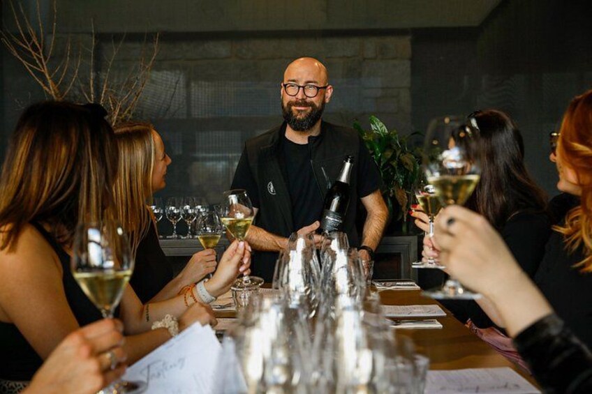 A group of people seated around a table with wine glasses, a man standing in the center, and a plant in the background. 3