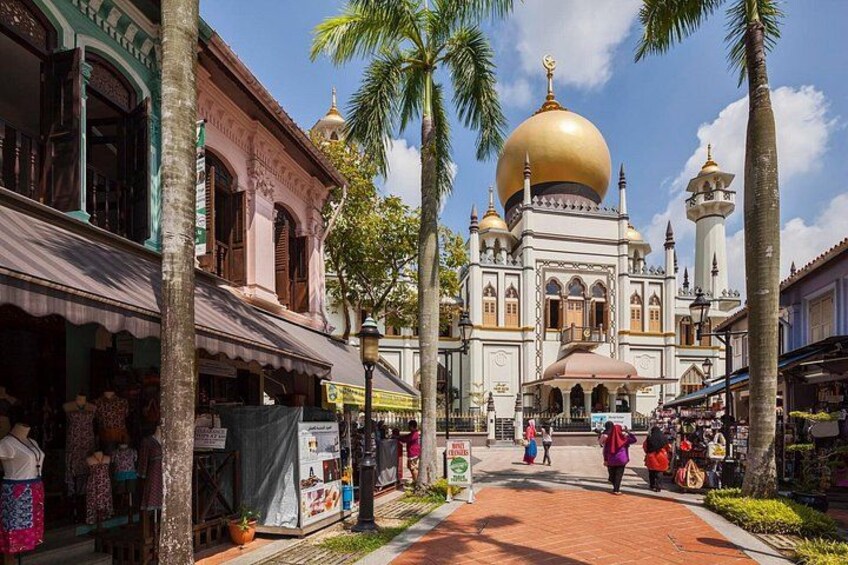 Eine Straße mit einer großen Moschee, deren Kuppel in Gold erstrahlt, Palmen und traditionellen Gebäuden. 1