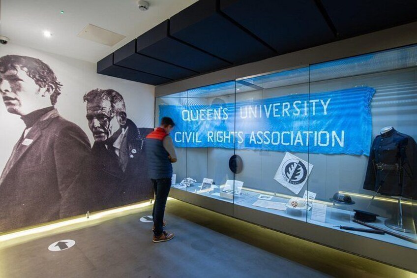 A museum exhibit with a large banner reading 'Queens University Civil Rights Association' and a display case with various items. 3