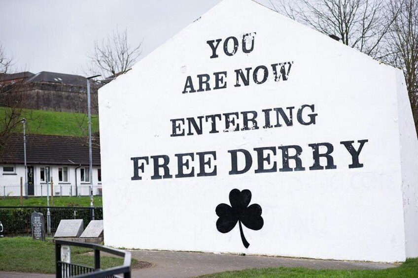 A large white sign with black lettering stating 'YOU ARE NOW ENTERING FREE DERRY' and a black clover symbol. 4