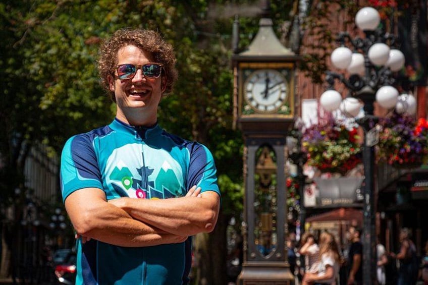 A person in a blue cycling jersey with arms crossed, standing in front of a clock tower with flowers. 1