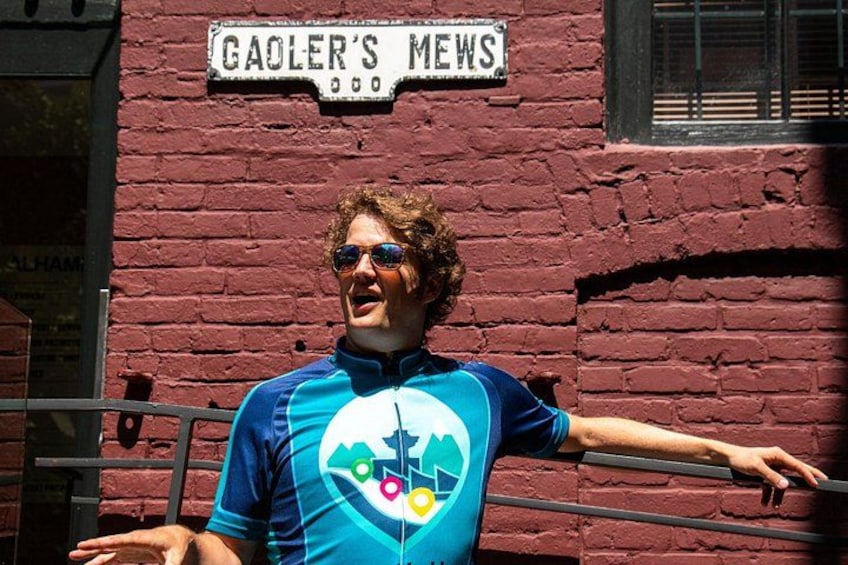 A person in a blue cycling jersey with a colorful logo, standing in front of a red brick building with a sign that reads 'GAOLER'S Mews.' 5