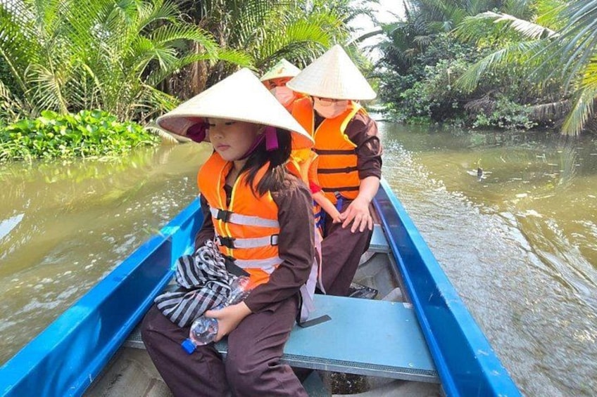 Two people in a boat wearing life jackets and conical hats, surrounded by water and greenery. 2