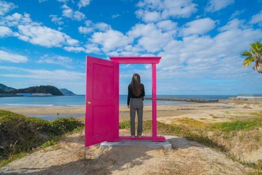 En person står framför en rosa byggnad på en strand, med en klar himmel och avlägsna landmärken i bakgrunden. 2